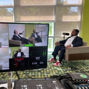 Man with headphones and a large microphone being interviewed by co-hosts shown on the video monitor at an event. 